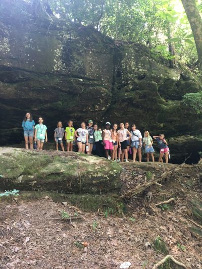 Campers lined up beneath a large mossy rock overhang on a hike