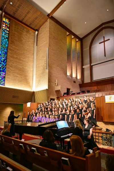 Wide view of the choir performing in a sanctuary with tall stained glass windows