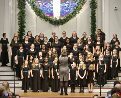 The full choir performing on stage beneath a stained glass window hung with holiday garland